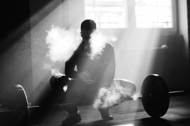 Black and white photo of muscular build man using sports chalk om his hands before exercising with a barbell in a gym. 