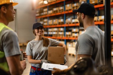 Happy industrial worker with cardboard box communicating with her colleagues in a warehouse. 
