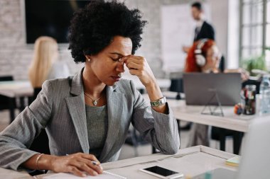 Tired black businesswoman feeling pain after a long day at work. There are people in the background. 