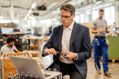 Quality control inspector measuring steel bolt with a caliper while using laptop in factory plant.  There are people in the background. 
