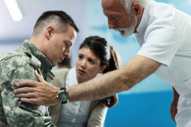 Mature doctor talking to army soldier who came with his wife to medical appointment at hospital. Focus is on soldier. 