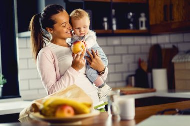Happy mother giving an apple to her baby son in the kitchen.