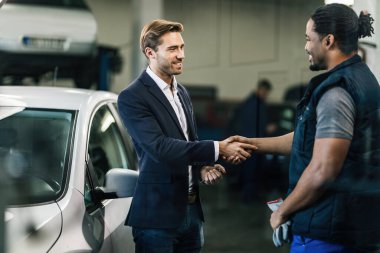 Happy manager and African American car mechanic handshaking in auto repair shop. 