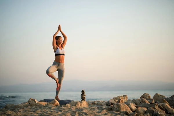 Athletic woman with hands above head practicing Yoga in tree pose on a beach rock at sunrise. Copy space.