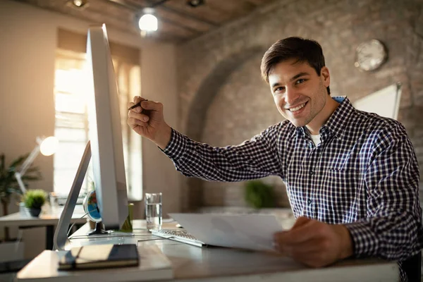 Young happy entrepreneur going through paperwork while using desktop PC in the office and looking at camera.