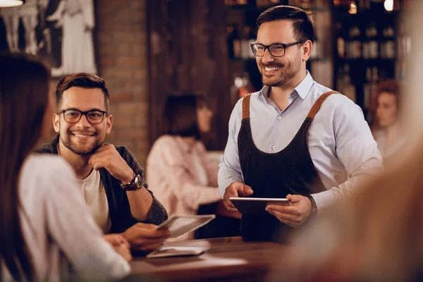 Happy waiter communicating with guests while taking their order on digital tablet in a pub.