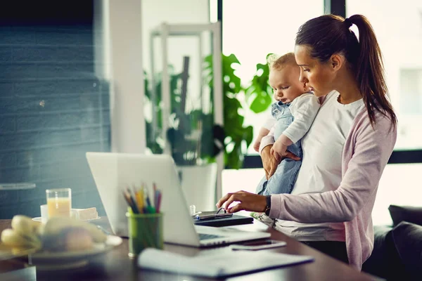 Young working mother using computer while holding her baby son at home.