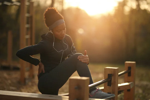 Black sportswoman feeling pain after injuring her knee during sports training in the park at sunset.