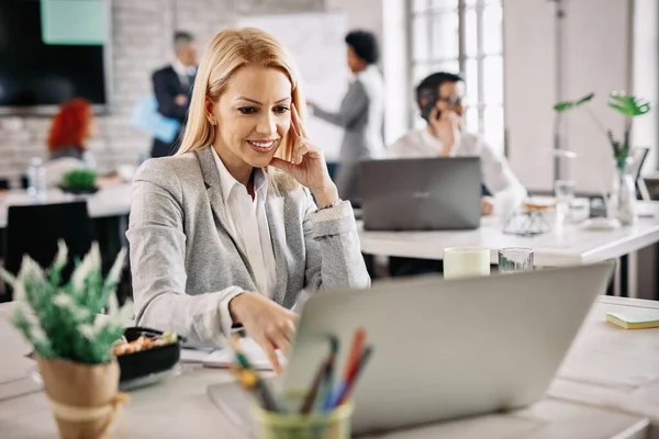 Happy mid adult businesswoman using computer and surfing on the Internet while working in the office. There are people in the background. 