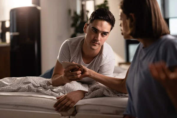 Young couple having relationship problems and arguing over mobile phone in the bedroom. Focus is on man.