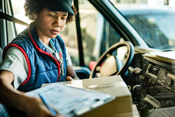 Black female courier getting ready for delivery and checking packages while sitting in delivery van.