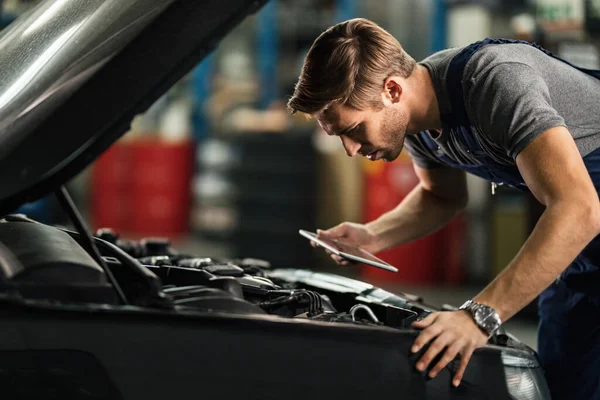 Young auto mechanic with touchpad examining car engine in repair shop.