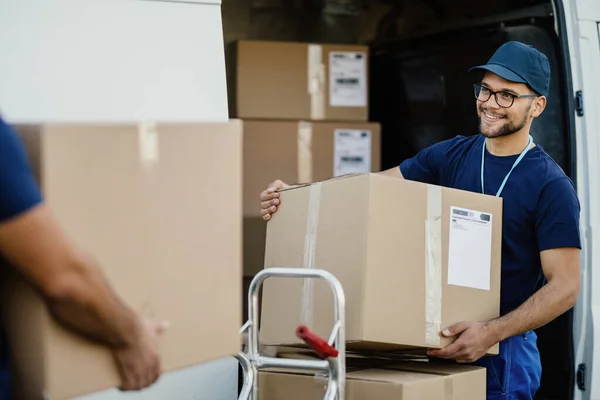 Young happy delivery man unloading boxes from a mini van and talking with his coworker. 
