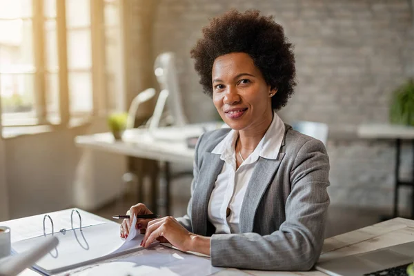 Black smiling businesswoman analyzing financial reports while working in the office and looking at the camera.