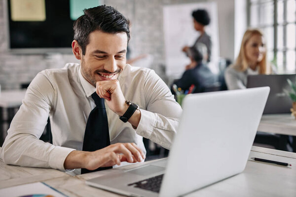 Smiling businessman sitting at desk in the office and working on laptop. There are people in the background. 