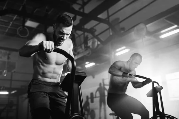 Black and white photo of athletic men having cross training on exercise bikes in a gym.