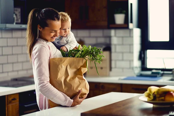 Happy mother with baby son placing groceries bag on kitchen counter.