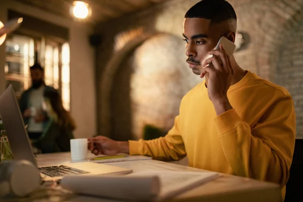 Young black entrepreneur working late on a computer and communicating on cell phone in the office.