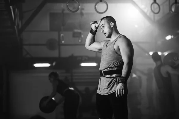 Black and white photo of muscular build man on a break during weight training in health club. There are people in the background.