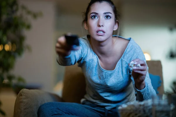 Young woman sitting in the dark and changing channels on TV while eating popcorn at home.
