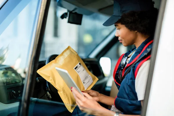 African American delivery woman using digital tablet while sitting in a van and checking package for the delivery.