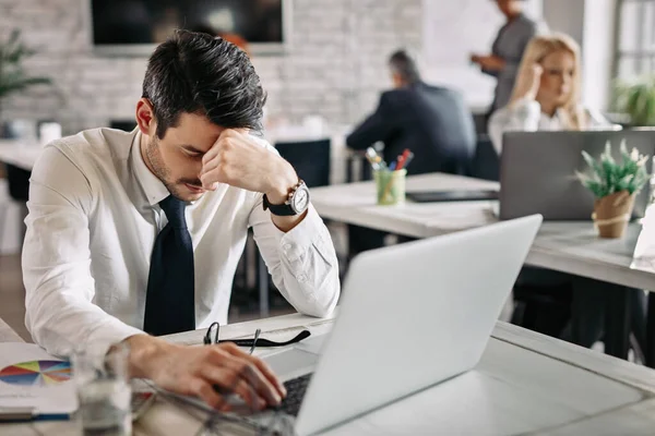 Tired businessman holding his head in pain while working on laptop and feeling exhausted from work. There are people in the background. 
