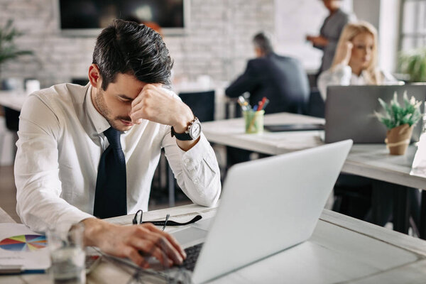 Tired businessman holding his head in pain while working on laptop and feeling exhausted from work. There are people in the background. 