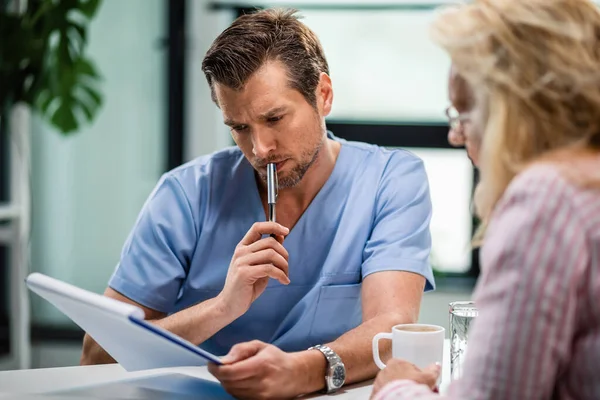 Serious Doctor Thinking While Going Medical Documents His Female Patient — Stockfoto