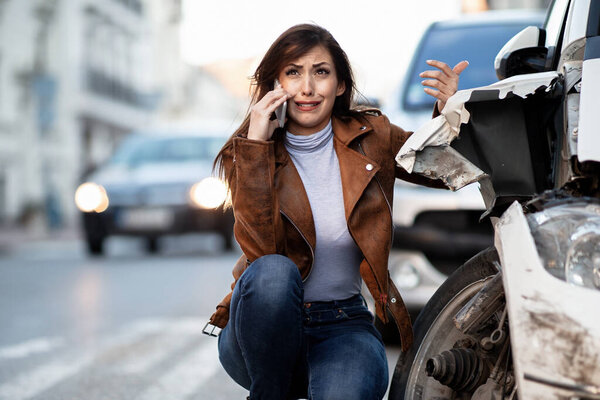 Crying woman talking on mobile phone and calling for road assistance after a car accident. 