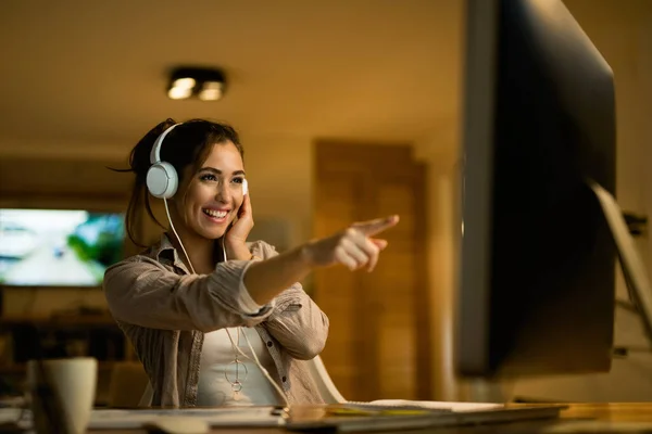 Happy woman with headphones pointing at computer screen while surfing the net in the evening at home.