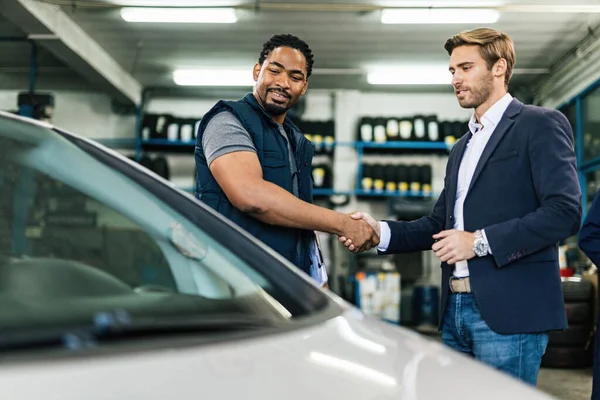 Happy black car repairman and young businessman handshaking in auto repair shop.