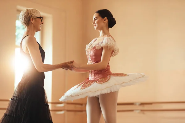 Happy mature ballet teacher and young ballerina talking while holding hands after rehearsal in ballet studio.