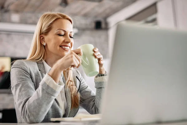 Smiling businesswoman taking a break and enjoying in cup of coffee while sitting at her desk in the office.