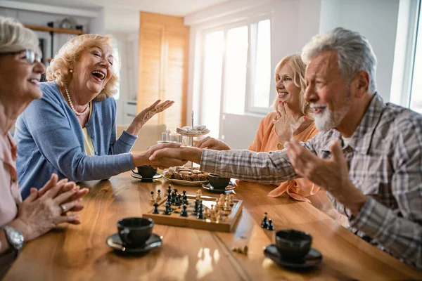 Group of happy seniors playing chess at home. Two of them are shaking hands and congratulating each other.