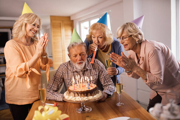 Group of happy senior friends celebrating Birthday at home. Man is blowing candles on a cake. 