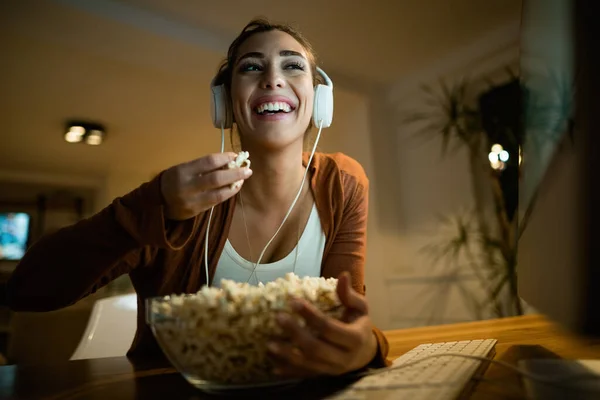 Low angle view of happy woman eating popcorn while using computer and watching something on the internet in the evening at home.