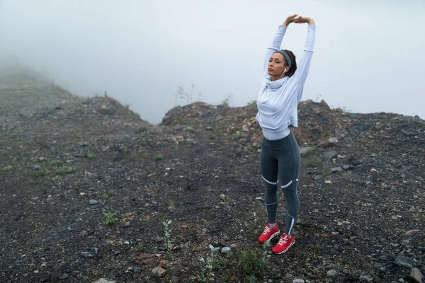 Active sportswoman preparing for morning run and stretching while standing near the cliff on cold misty weather. Copy space.  