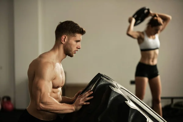 Young athletic man doing tire flip exercise on cross training in a gym. There is a woman working out with weights in the background.