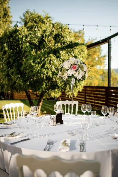 White dining table with floral centerpiece at outdoors wedding reception.