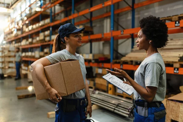 African American warehouse worker talking about delivery schedule with her coworker in industrial storage room.