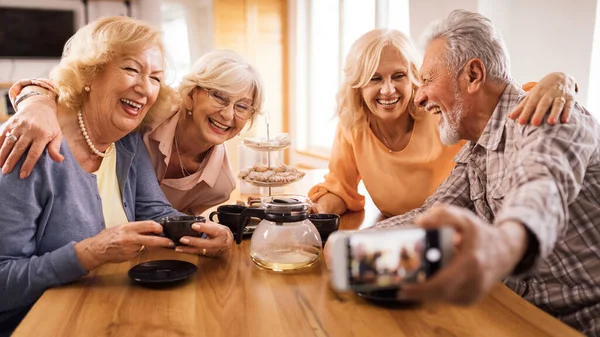 Group of happy senior people using mobile phone and taking selfie during tea time at home.