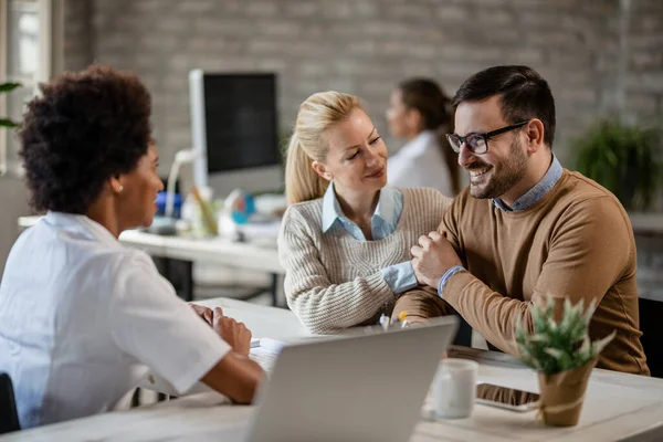 Young happy couple talking to African American doctor and feeling relieved  after hearing good news during medical appointment. Focus is on man. 