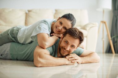 Happy husband and wife relaxing on the floor at home and looking at camera. Woman is lying on man's back.