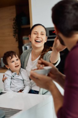 Happy family having fun during breakfast at dining table. Focus is on woman feeding her husband. 