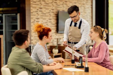 Young waiter communicating with guests and helping them to chose their order from a menu.