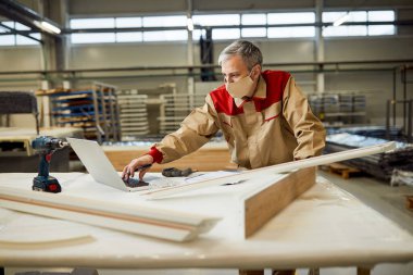 Manual worker using computer while working at carpentry workshop and wearing protective face mask due to coronavirus pandemic. 