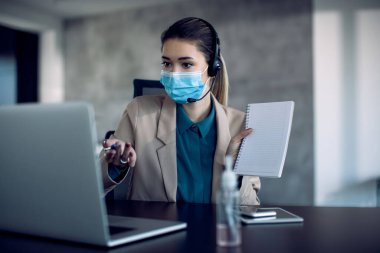 Young businesswoman explaining something during video call over laptop and wearing face mask due to coronavirus pandemic. 