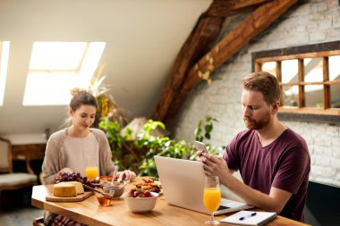 Redhead man using smart phone and laptop during breakfast with is girlfriend at dining table at home.