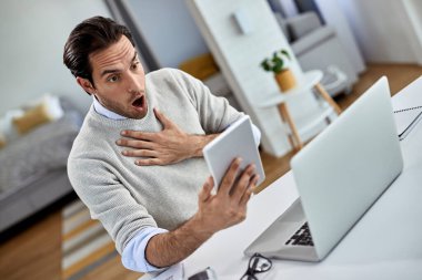 Shocked businessman using touchpad and reading something in disbelief while working at home. 