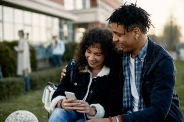 Happy African American student and his girlfriend using mobile phone and having fun outdoors at campus. 